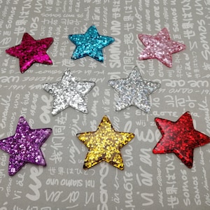 Puede incluir: Surtido de siete adornos de estrellas de purpurina en varios colores, incluyendo rosa, azul, plata, oro, rojo y morado. Las estrellas son todas del mismo tamaño y forma, y están dispuestas en un patrón aleatorio sobre un fondo gris.