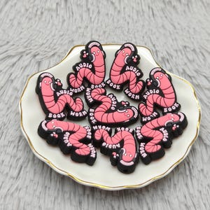 Puede incluir: Un conjunto de dijes para zapatos rosas y negros con forma de gusanos de dibujos animados, cada uno con auriculares y la etiqueta "Audio Book Worm". Los dijes están dispuestos sobre un plato blanco con borde dorado, sobre un fondo suave y texturizado.