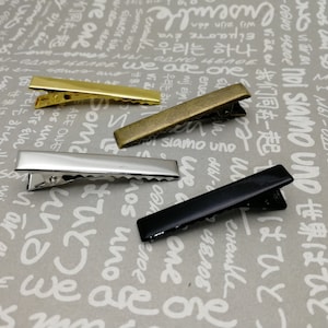May include: Four hair clips in different colours: gold, silver, bronze, and black. The clips are all the same size and shape, with a rectangular metal body and a spring-loaded clasp.