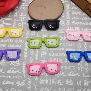 Puede incluir: Seis pares de gafas de sol pixeladas de colores con monturas negras. Las gafas de sol son de plástico y tienen un diseño pixelado.