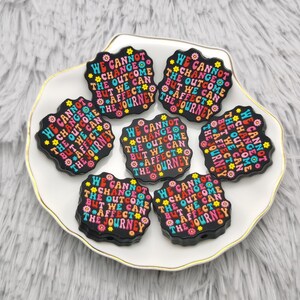 May include: Seven black square-shaped buttons with a colorful floral design and the text "We cannot change the outcome but we can affect the journey" repeated on each button.