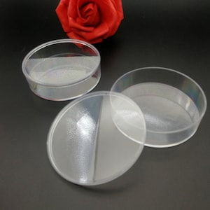 Op de afbeelding: Drie heldere, ronde plastic containers met deksels. De containers zijn transparant, waardoor de inhoud zichtbaar is. De deksels zijn licht ondoorzichtig. De containers staan op een donkere ondergrond.