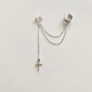 May include: Silver ear cuff with a dangling chain and a small cross charm. The chain is attached to a stud earring.