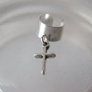 Puede incluir: Un pendiente de aro de plata con un dije de cruz colgante. El aro es de metal y tiene un diseño simple. El dije de cruz también es de metal y tiene un diseño simple.