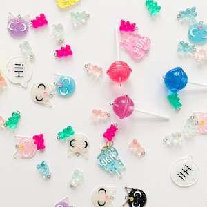 Op de afbeelding: Assortiment van kleurrijke harscharms, waaronder gummyberen, katten, lollies en spraakballonnen. De charms zijn glitterend en doorschijnend. De charms zijn perfect voor het maken van sieraden, sleutelhangers en andere knutselprojecten.