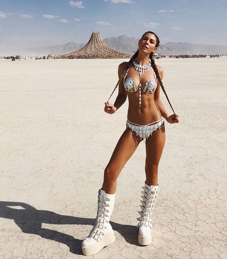 May include: A woman wearing a white, sequined bikini top and matching bottoms with a white, beaded necklace. She is wearing white, platform boots with multiple buckles. The woman is standing in a desert landscape.