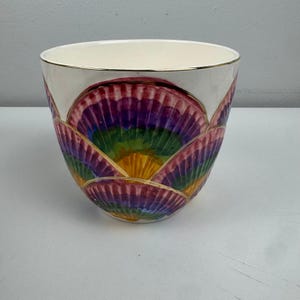 May include: A white ceramic bowl with a gold rim. The exterior features a colorful, fan-like design in shades of purple, green, yellow, and red. The bowl's design is reminiscent of a flower or seashell.