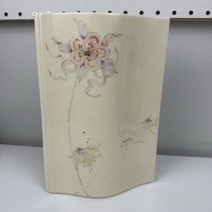 May include: A cream-colored ceramic vase with a floral design. The vase features a large, detailed flower in shades of pink and purple, with a yellow center. Additional smaller flowers and gold accents are also present.