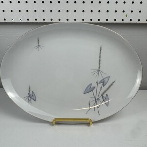 May include: Oval white serving platter with a silver rim and a gold-colored metal stand. The platter features a delicate design of blue and gray stylized plants. A vintage piece for serving food.