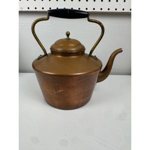 May include: A copper tea kettle with a black handle and brass accents. The kettle has a rounded body, a spout, and a lid with a small knob. The handle is arched, and the kettle appears to be vintage.