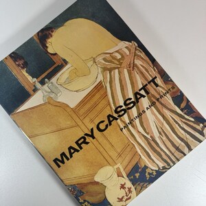 May include: A book cover featuring a painting by Mary Cassatt. The artwork depicts a person washing at a sink, with a striped robe. The title "MARY CASSATT PAINTINGS AND PRINTS" is printed in large black letters.