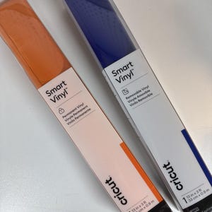 Puede incluir: Dos rollos de vinilo inteligente Cricut en naranja y azul. El rollo naranja está etiquetado como "Vinilo Permanente" y el azul como "Vinilo Removible". Cada rollo mide 33 cm por 0,9 m.