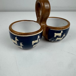 Op de afbeelding: Een dubbele keramische schaal met een centrale boogvormige handgreep. De buitenkant is donkerblauw met handgeschilderde witte lama's en de tekst MAKY CUSCO. De binnenkant van beide kommen is wit, en de rand en handgreep zijn van bruine klei.