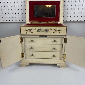 May include: A cream-colored wooden jewelry box with a hinged lid, an interior mirror framed in red velvet, and two side doors. The front features one top drawer with a decorative metal handle and three lower drawers with small round knobs.