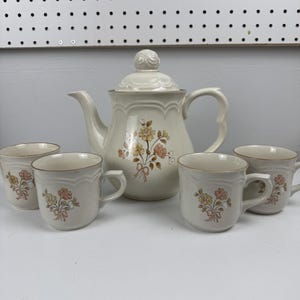 May include: Cream-colored ceramic tea set featuring a teapot with a lid and four matching mugs. The teapot and mugs are adorned with a floral design in shades of yellow, peach, and brown. The set has a classic, vintage aesthetic.