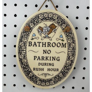 Puede incluir: Letrero de baño ovalado de cerámica con fondo crema y borde floral negro. El letrero presenta un oso en una bañera y el texto "BATHROOM NO PARKING DURING RUSH HOUR". Dos pequeños acentos decorativos marrones están en la parte superior.