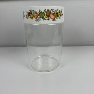 May include: A clear glass canister featuring a white band around the top rim. The white band is decorated with a colourful botanical pattern of various fruits, vegetables, and small flowers in shades of orange, green, brown, and yellow.