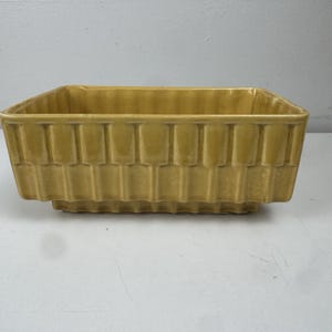 Op de afbeelding: Een rechthoekige, mosterdgele keramische plantenbak met een getextureerd, verticaal patroon. De plantenbak heeft een iets verhoogde basis en een vierkante bovenkant, geschikt voor binnen of buiten. Het algehele ontwerp is eenvoudig en klassiek.
