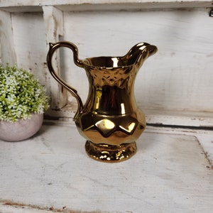 May include: A gold ceramic pitcher with a textured surface and a scalloped rim. The pitcher has a handle and a wide base.