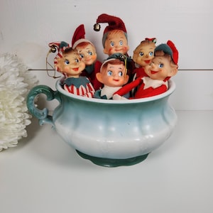 May include: A group of six vintage elf ornaments in a blue and white ceramic pitcher. The elves are wearing red and green clothing and have white faces with painted features.