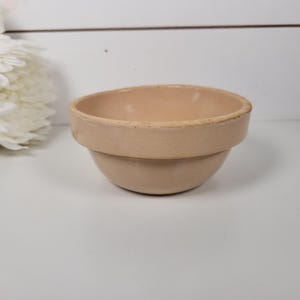 May include: A beige ceramic bowl with a rounded base and a wider, stepped rim. The bowl is set against a white background with a cluster of white flowers to the left. The bowl is likely for kitchen or home decor.