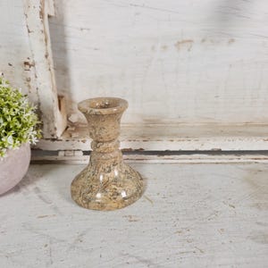 May include: A beige stone candlestick holder with a textured surface. The candlestick has a wide base, a decorative middle section, and a flared top. A small, round, pink pot with a green and white plant is to the left.