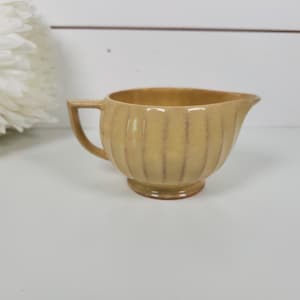 May include: A small, light yellow ceramic creamer pitcher featuring a ribbed, vertical fluted pattern along its rounded body. The pitcher has a single angular handle on one side and a small pouring spout, finished with a glossy glaze.