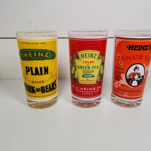 May include: Three vintage glass tumblers with Heinz soup labels. The first tumbler is yellow with the text "Heinz Plain Baked Pork and Beans". The second tumbler is red with the text "Heinz Cream of Green Pea Soup". The third tumbler is red with the text "Heinz's Tomato Soup".