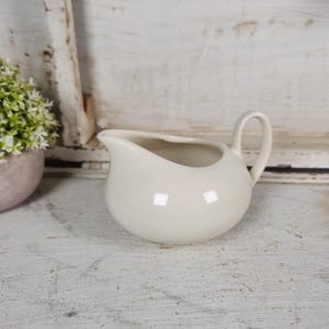 May include: Cream-colored ceramic creamer with a rounded body and a curved handle. The creamer has a spout for easy pouring. A small, light pink pot with a green plant is visible in the background.