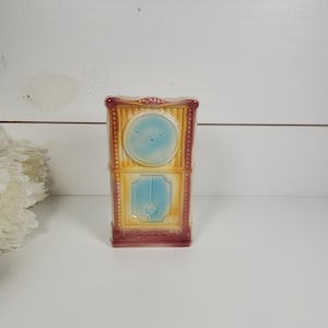 May include: A vintage ceramic clock figurine with a yellow and blue painted design. The clock has a red frame and a white face with black hands.
