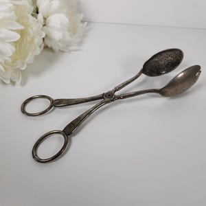 May include: Silver salad tongs with a decorative design. The tongs have two oval-shaped ends.