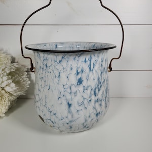 May include: A white and blue marbled enamel bucket with a dark rim and a thin, arched metal handle. The bucket features a speckled, mottled pattern throughout its exterior surface, with a smooth, rounded base and a wide, open top opening.