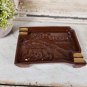May include: Brown ceramic ashtray shaped like the state of South Dakota with gold accents. The ashtray features the names of cities in South Dakota, including Spearfish, Rapid City, Pierre, and Wall.