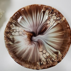 May include: A decorative ceramic plate with a unique design. The plate features a brown and white color scheme with a swirling pattern. The rim of the plate has a textured, raised design. The plate is likely used for serving or display.