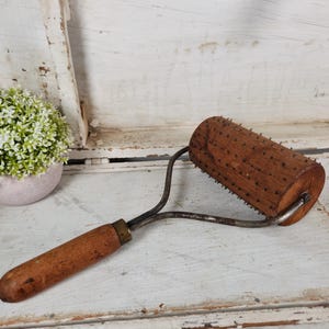 May include: An antique wooden rolling pin with metal hardware. The roller is cylindrical with small, evenly spaced wooden pegs. The handle is made of wood and is attached to the roller with a curved metal frame.