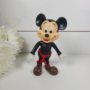 Puede incluir: Una figura vintage de Mickey Mouse de pl&aacute;stico negro con tela roja alrededor de la cintura. La figura tiene una cara sonriente con ojos negros y una nariz roja.