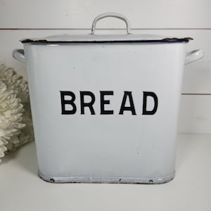 May include: White enamel bread box with a lid and handle. The word "BREAD" is printed in large black letters on the front. The box has a rectangular shape with rounded corners and two side handles. A classic kitchen storage container.