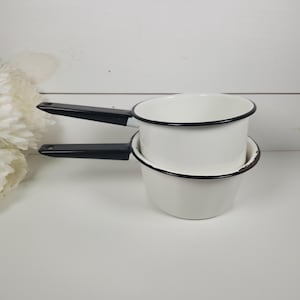 May include: Two white enamelware saucepans with black handles and rims are stacked on a white surface. The pans have a classic design, with a rounded shape and a single handle. The background includes white flowers and a white wall.