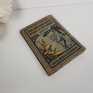 May include: An antique book titled "The Progressive Road to Reading Book One" with a faded green cover. The cover features illustrations of a person and yellow flowers. The book is from Silver, Burdett & Company.