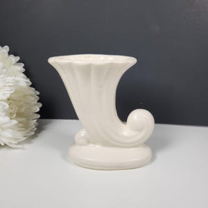May include: Cream-colored ceramic cornucopia vase. The vase has a curved, horn-like shape with a flared opening and a rounded base. The design includes a decorative swirl and vertical ridges. A white flower is partially visible on the left.