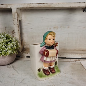 May include: A white ceramic planter shaped like a girl holding an accordion. The girl is wearing a pink dress, a blue headscarf, and brown shoes. The planter has green grass and brown tree stumps at the bottom.