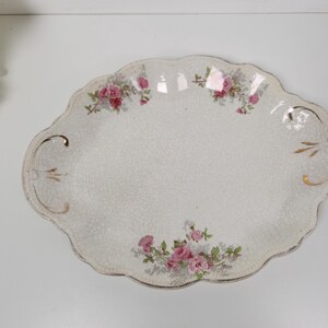 May include: An oval, antique serving platter with a scalloped edge. The white ceramic platter features pink rose floral designs and gold accents. The platter has a crackle finish, adding to its vintage charm.