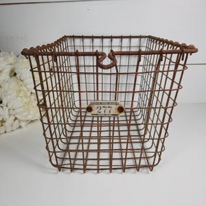 May include: A rusty brown wire basket with a rectangular shape. The basket has a metal tag that reads "RASPE WIRE WORKS 277". The basket has a handle and is likely used for storage or display.