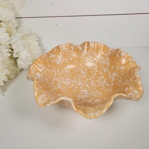 May include: A decorative, ruffled-edge bowl with a speckled, light orange and white design. The bowl is made of ceramic or glass and is set against a white background with white flowers.