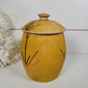 May include: A yellow ceramic jar with a lid. The jar has a rounded shape and is decorated with black and red painted lines. The lid has a small knob on top. The jar is sitting on a white surface.