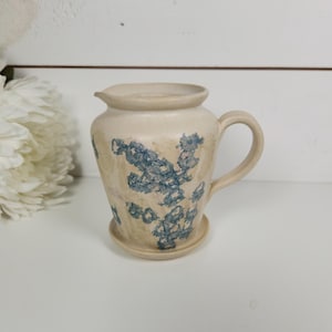 May include: A beige ceramic pitcher with a handle and a small spout. The pitcher is decorated with blue floral patterns. It sits on a small, attached saucer. The pitcher is likely handmade and suitable for serving beverages.