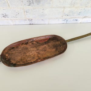 May include: An oval-shaped copper pan with a dark, weathered patina and a textured surface. The pan features a long, flat metal handle extending from one side and a small circular metal ring attached to the opposite end, secured with rivets.