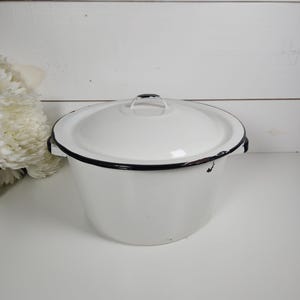 May include: White enamelware pot with a lid. The pot has a black rim and handle. The pot is sitting on a white surface. The background is a white wooden wall.