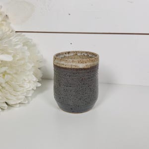 May include: A handmade ceramic tumbler with a speckled, dark gray body and a light beige rim. The cup has a textured surface and a rounded base. The background is a white surface with a white flower.