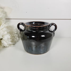May include: A small, dark-glazed ceramic vase with two loop handles. The vase has a rounded body and a glossy, reflective surface. The color is a deep, rich black with hints of blue and brown. The vase is likely handmade and suitable for home decor.